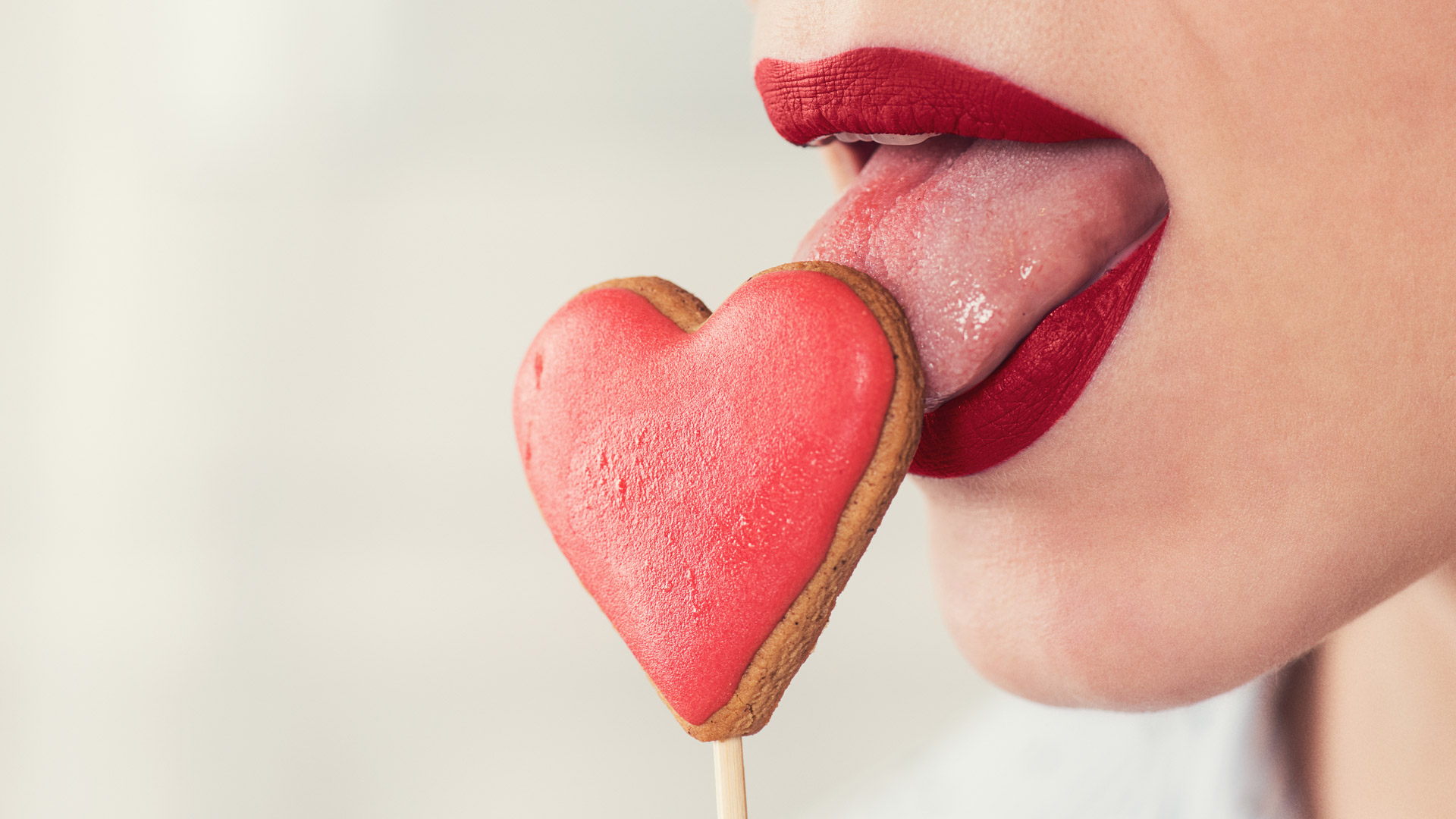 Female mouth with dark red lipstick licking a lollipop in the shape of a love heart.