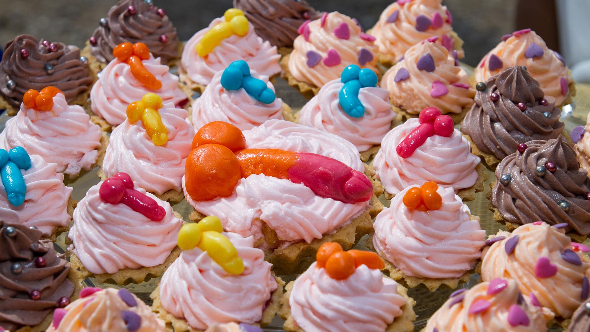 Many cupcakes of different colored toppings in the shapes of penises.