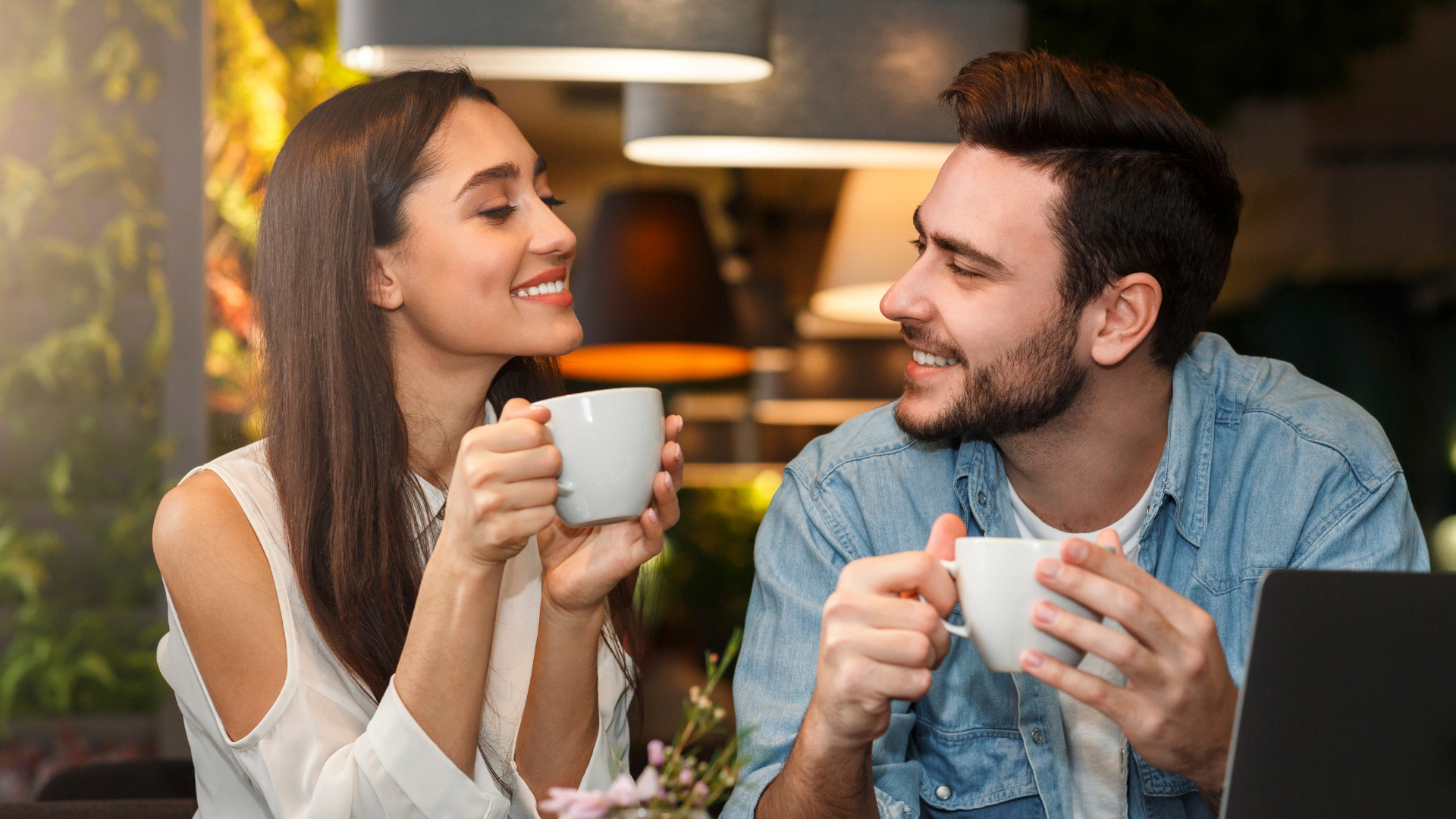 Brunette man and brunette woman sitting together having a coffee, and smiling at one another