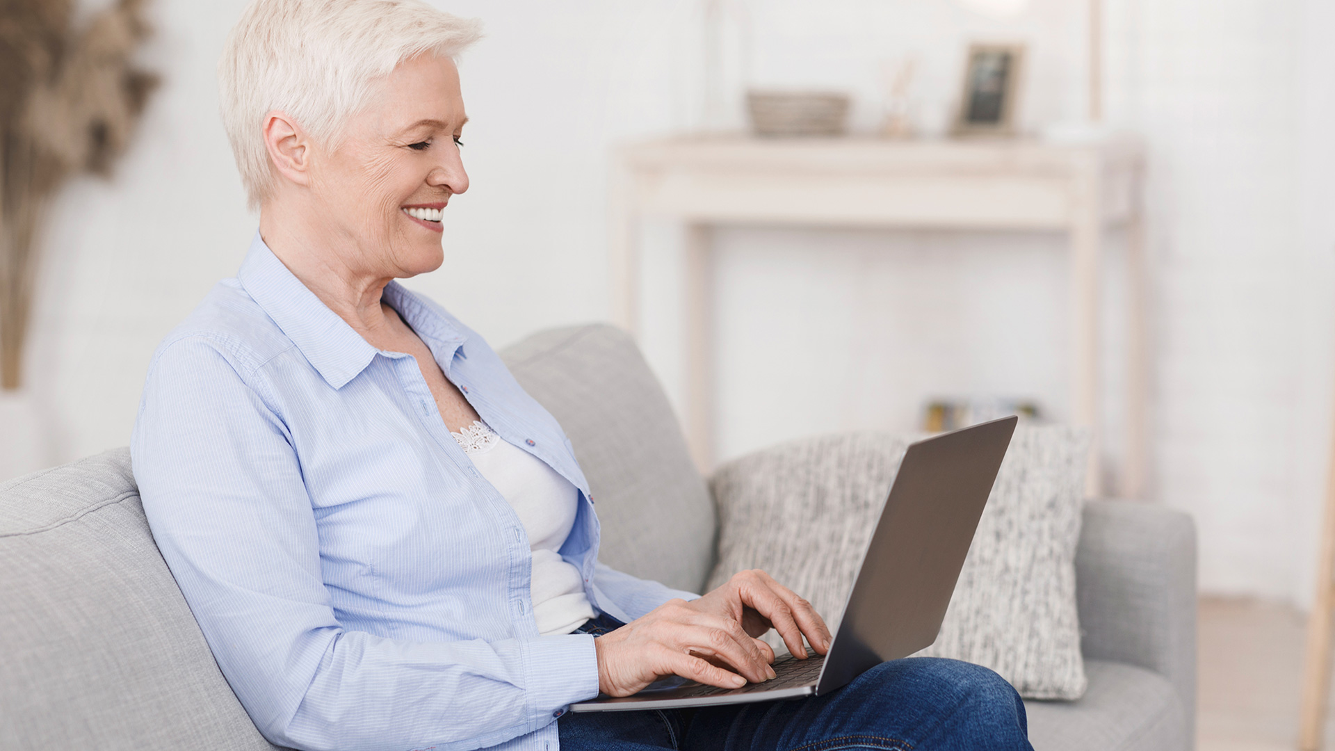 Older woman with short white hair and light blue shirt sitting down on couch with laptop on lap and smiling while typing.
