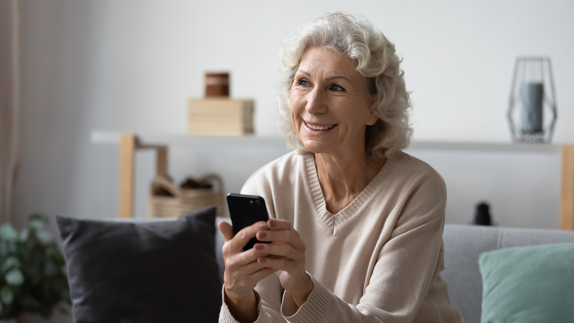 Older woman with white, medium-length hair holding her phone while smiling. She is sitting in her living room.