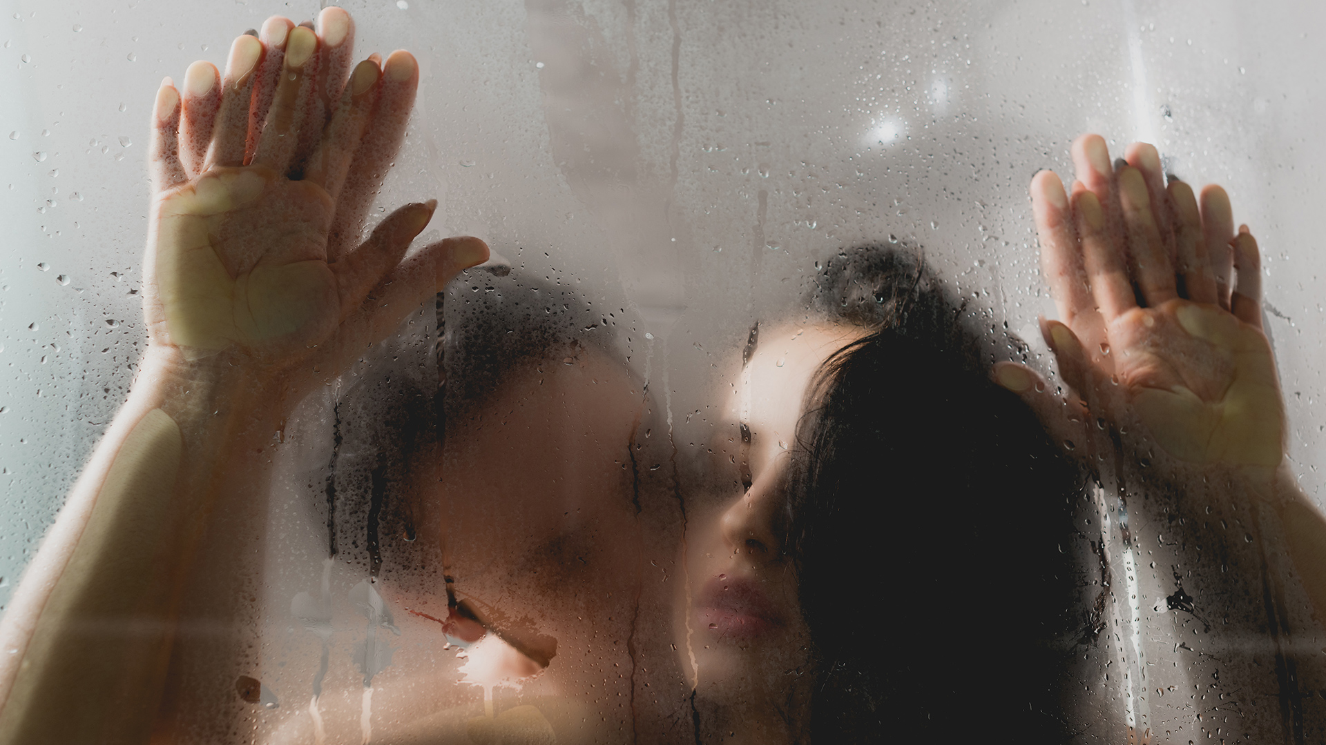 Woman being pinned against the shower glass door by a man behind her while holding her hands up and kissing her neck