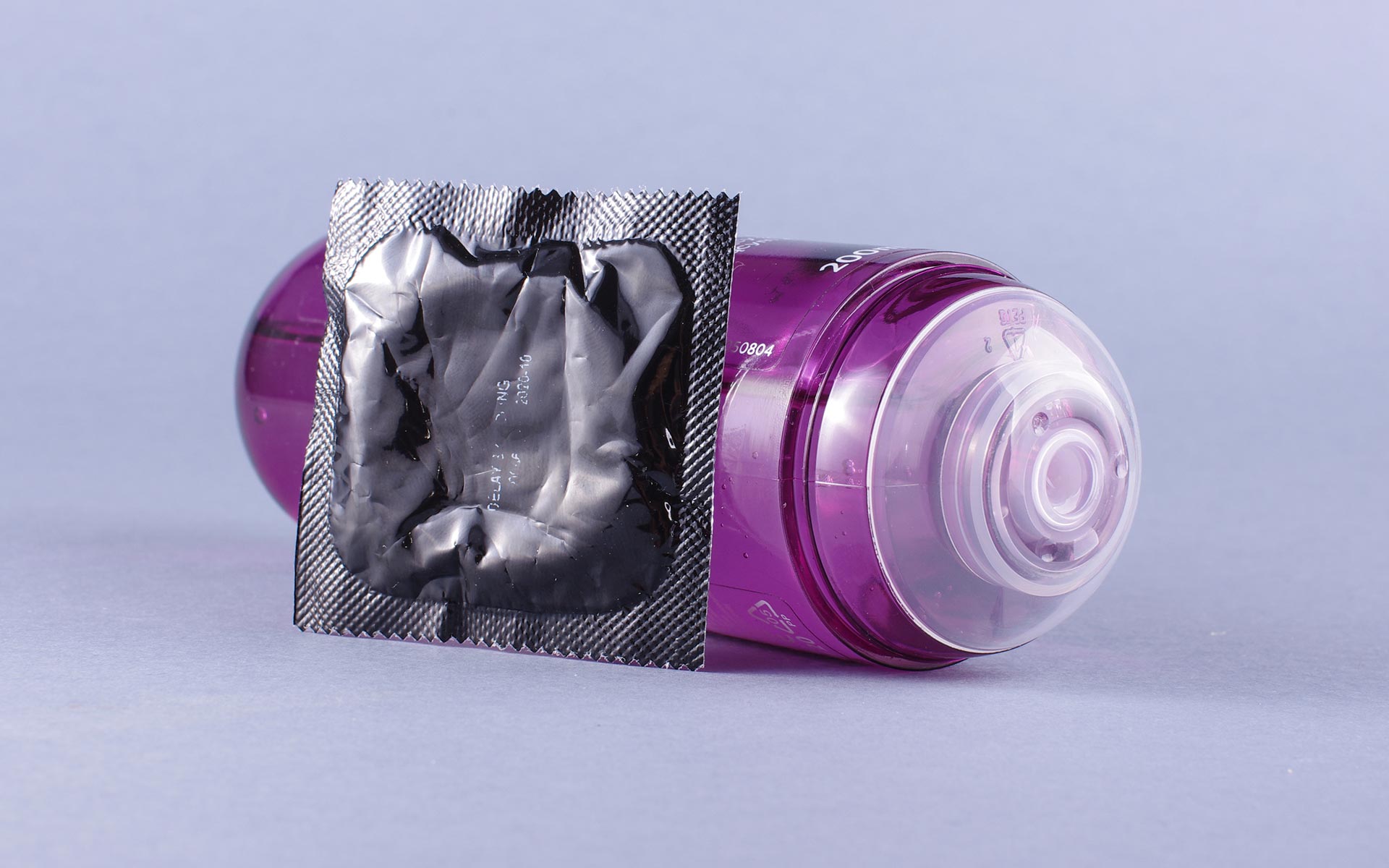 Purple bottle of lube on the ground with an unopened silver condom wrapper leaning against it from the front.