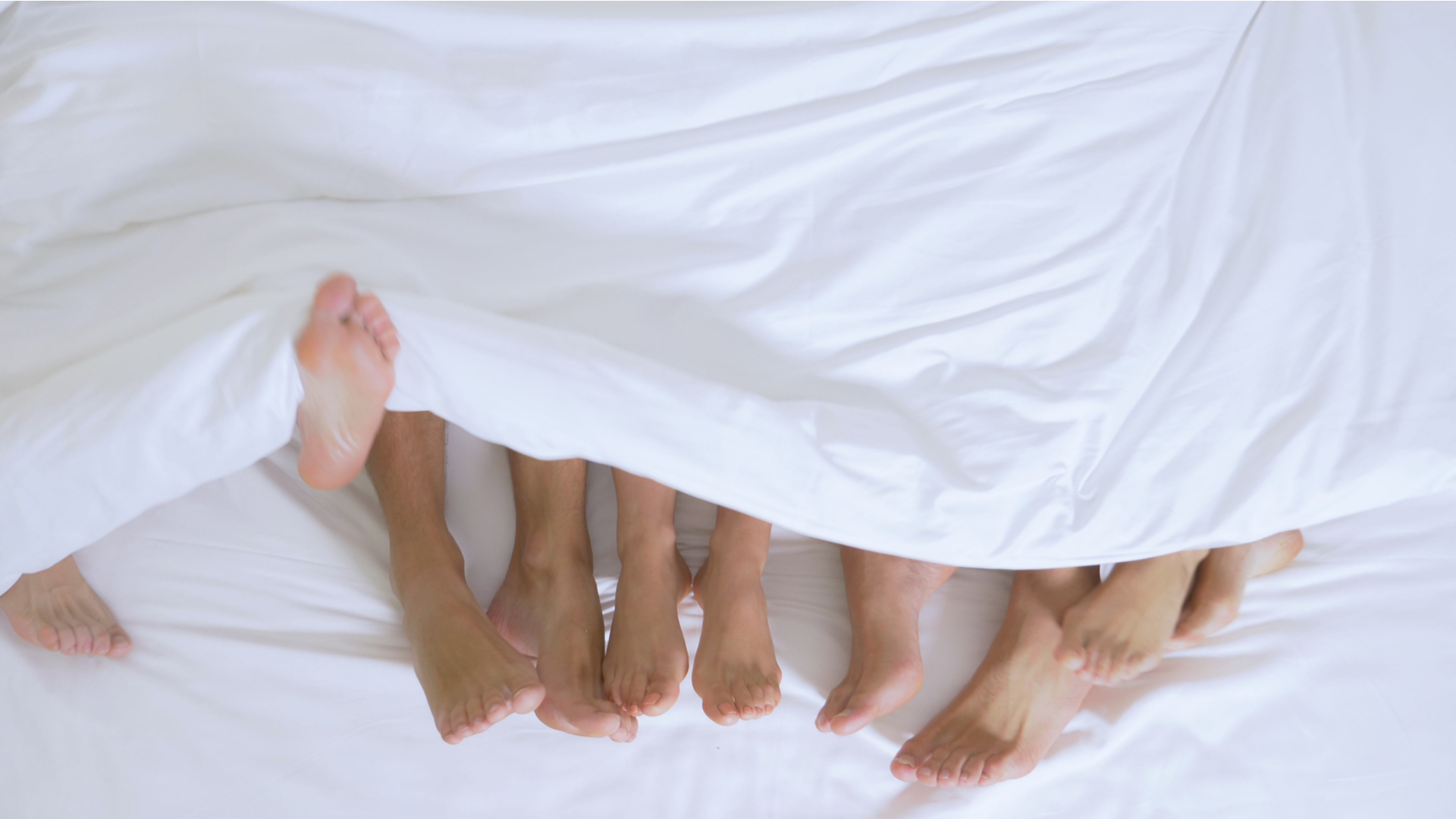 Feet of multiple people lying in the same bed, coming out of the bottom of a white duvet.