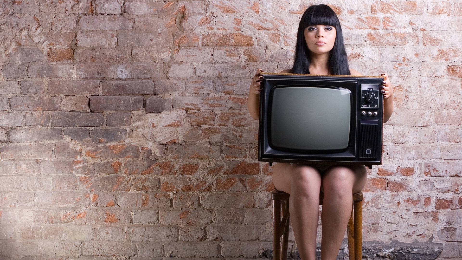 Naked black-haired woman sitting on a chair with an old TV on her lap covering her body. Brick wall background.